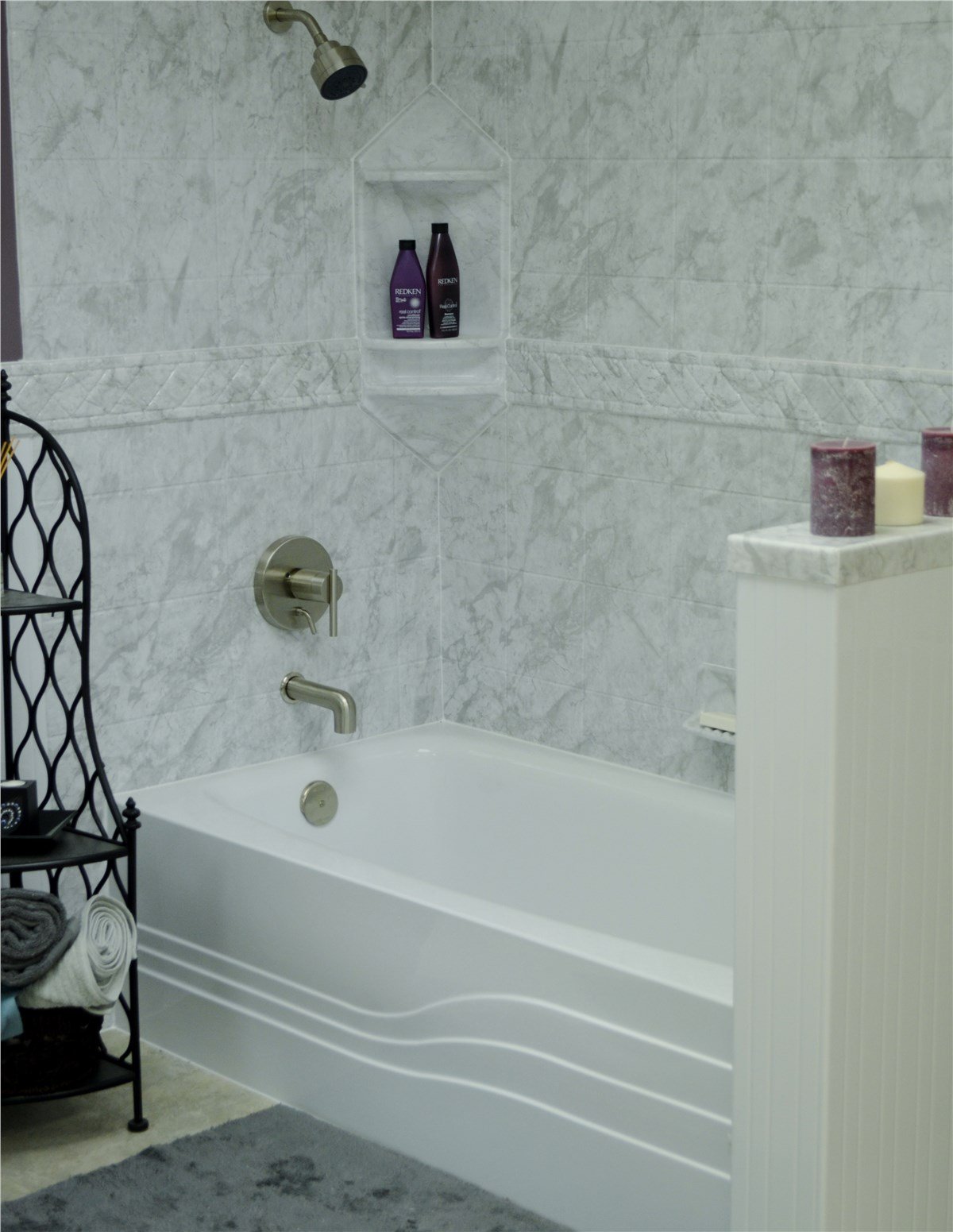 A modern bathroom with a marble-tiled bathtub, purple shampoo bottles, candles, and a black shelving unit.