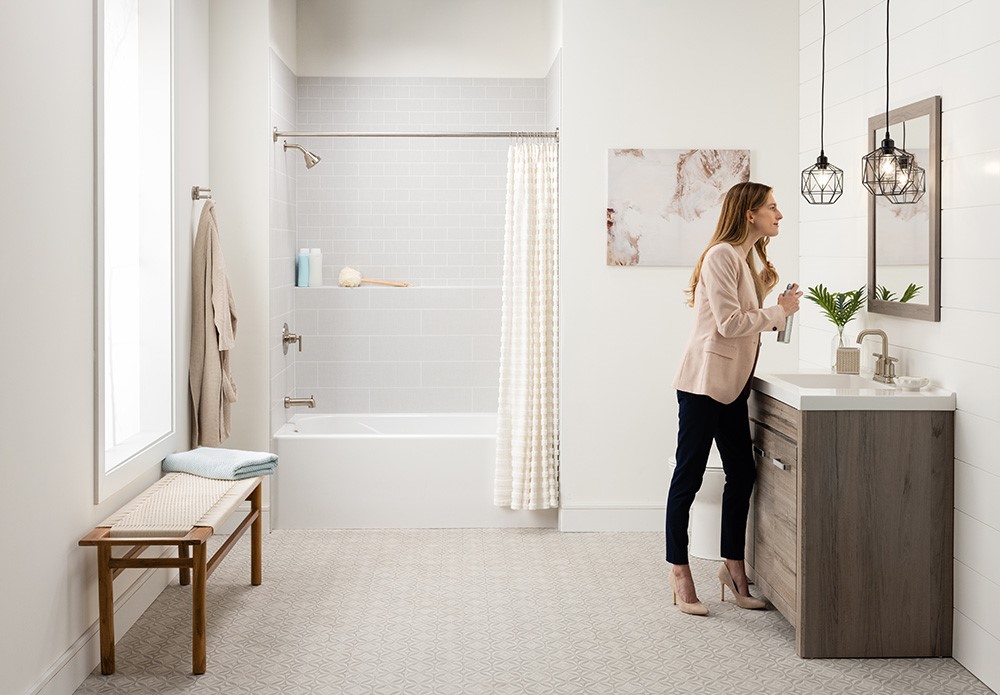A woman stands in a bathroom beside a sink and shower, with neutral-colored walls and soft lighting