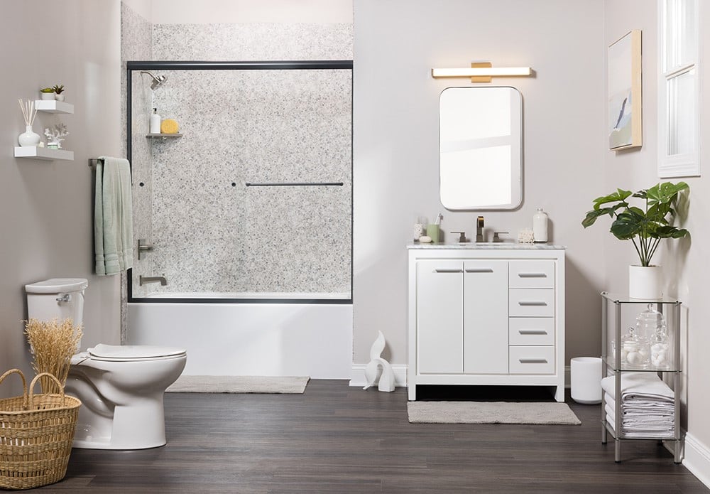 A modern bathroom with a speckled-tiled bathtub, glass sliding door, white vanity, and decorative shelving unit.