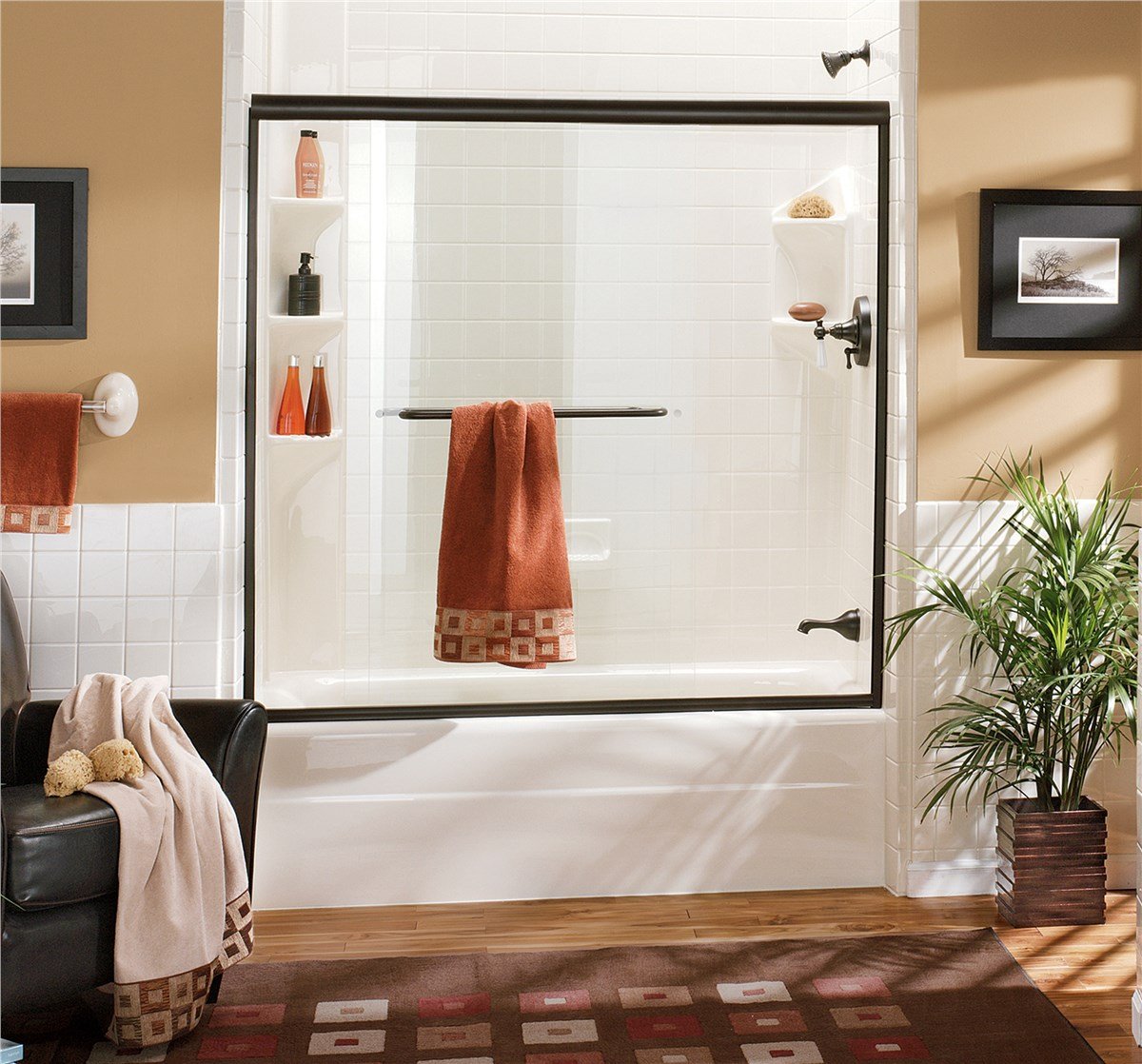 A bathroom with a white tiled bathtub and black-framed glass sliding door, coordinated rust-colored towels, and wood flooring.